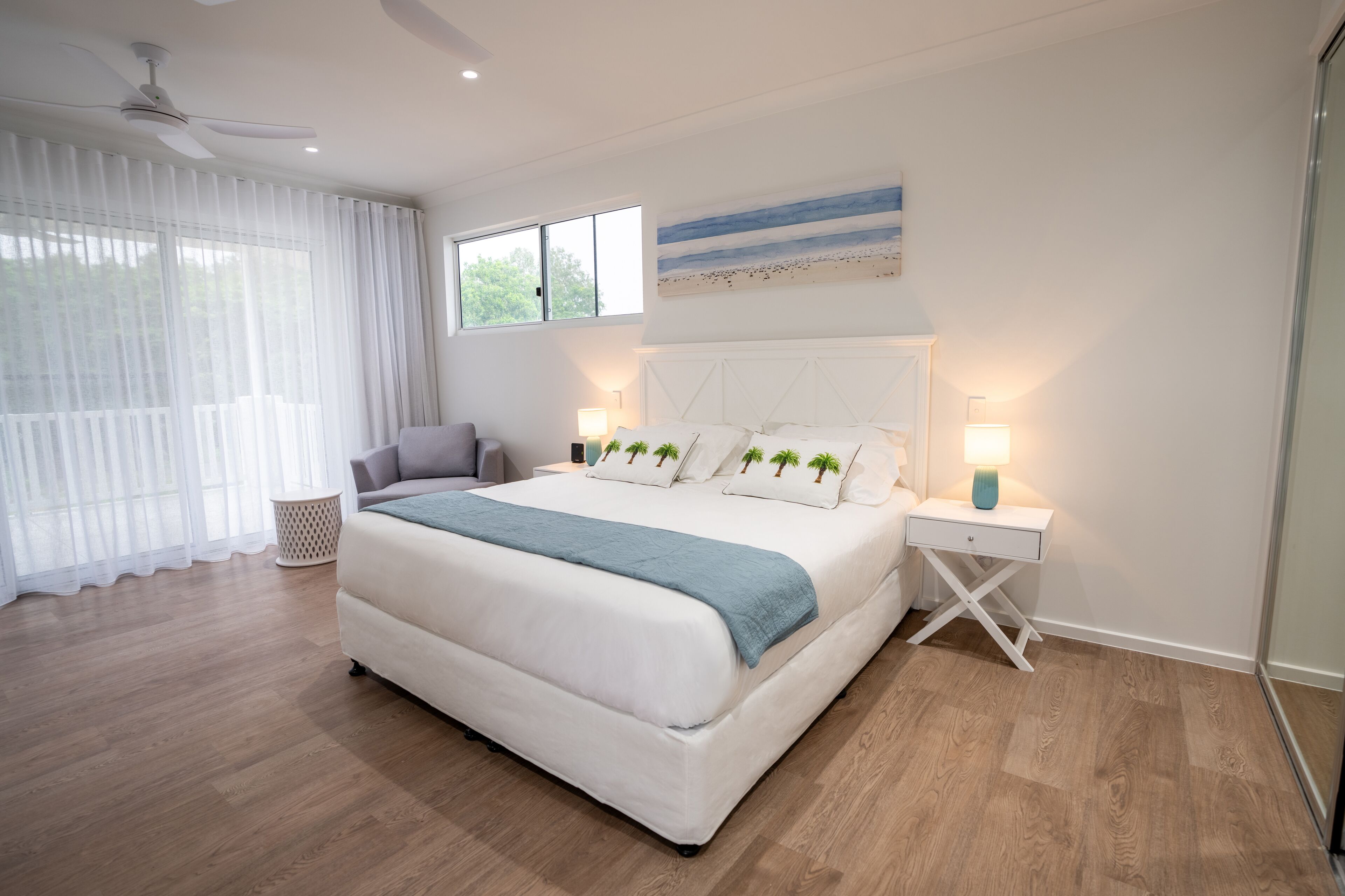 A modern bedroom with a large bed, a ceiling fan, a window with sheer curtains, and a wall-mounted artwork.