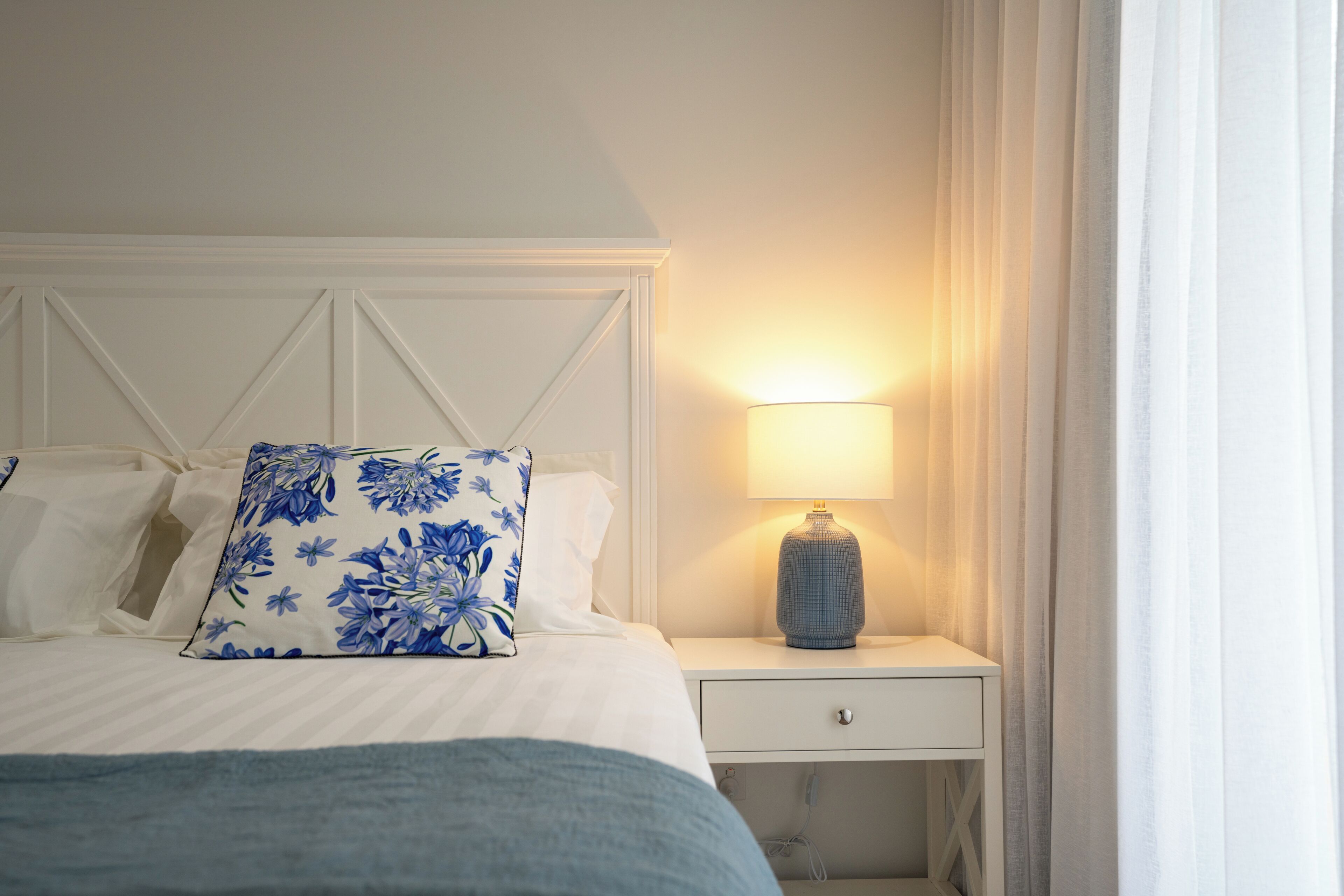 Una cama bien hecha con un cojín floral, una lámpara de noche y una mesita de noche blanca con un cajón.