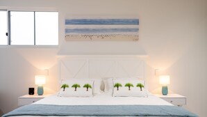 Ein Schlafzimmer mit einem Bett, zwei Nachttischen, einem Fenster und einem Wandbild zum Thema Strand.