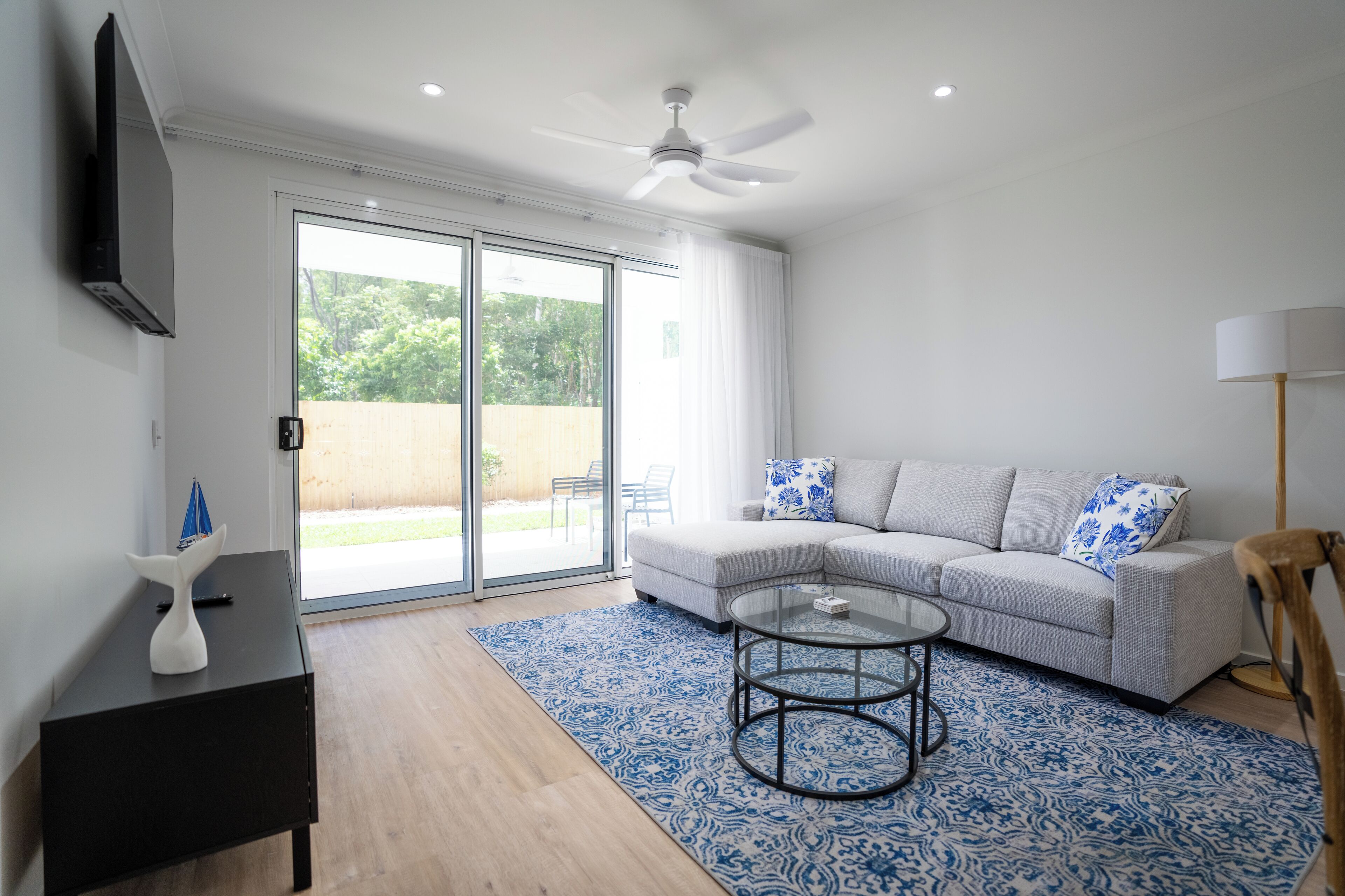 A modern living room with a grey sofa, a glass coffee table, a black side table, and a ceiling fan.