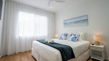 A neatly made bed with a white and blue floral pillow set, a dark blue throw blanket, and a white towel folded on the bed. There is a small bedside table with a lamp and a remote control on it. A ceiling fan is visible above the bed, and a painting of a beach scene hangs on the wall.