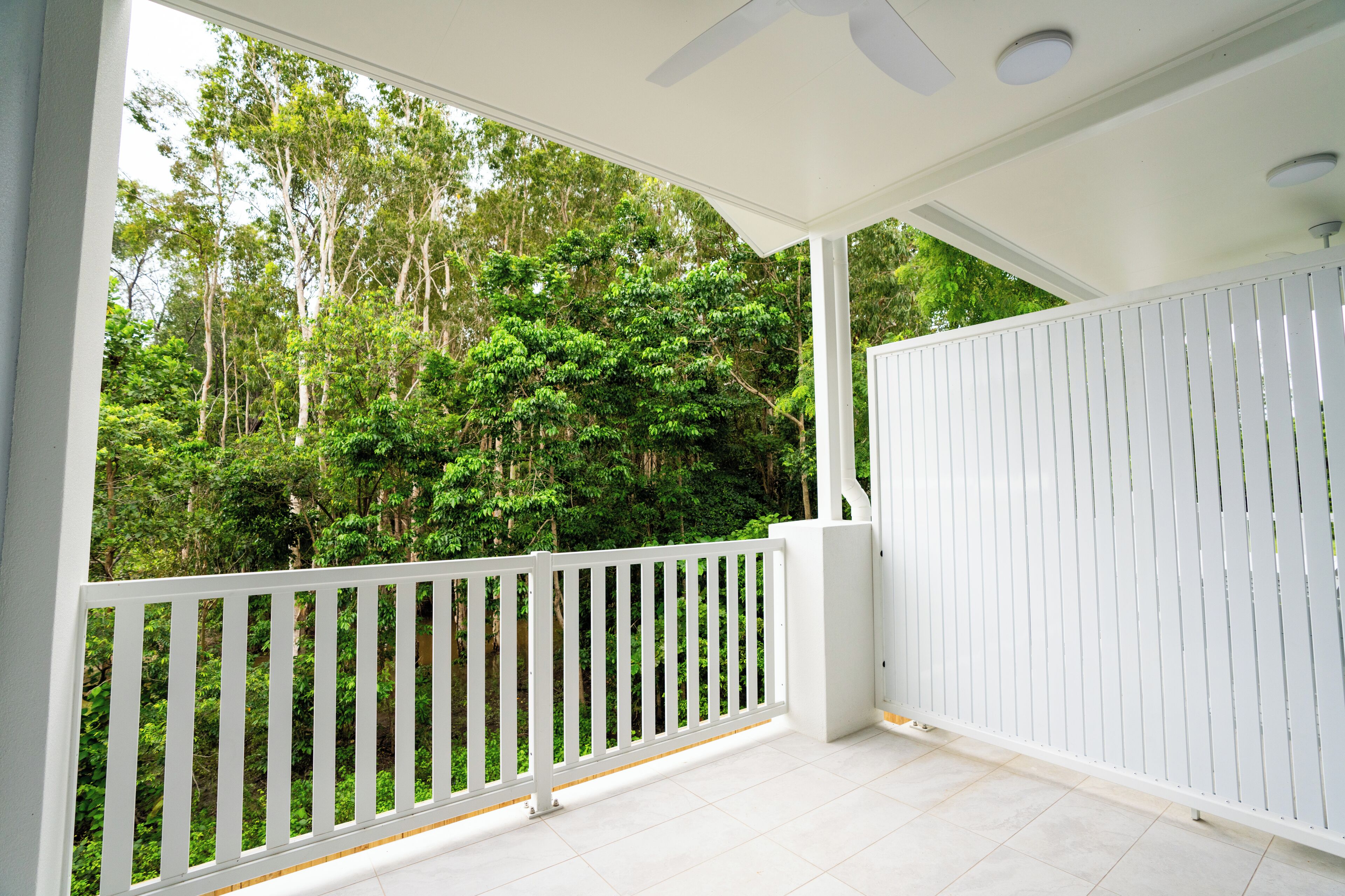 A covered balcony with white railings, a ceiling fan, and a view of a dense forest.