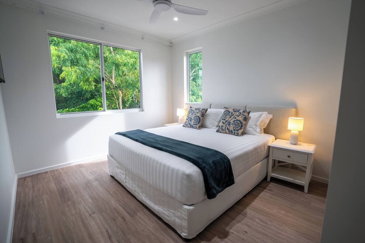 A bedroom with a large bed, two bedside tables, and two windows with a view of trees.