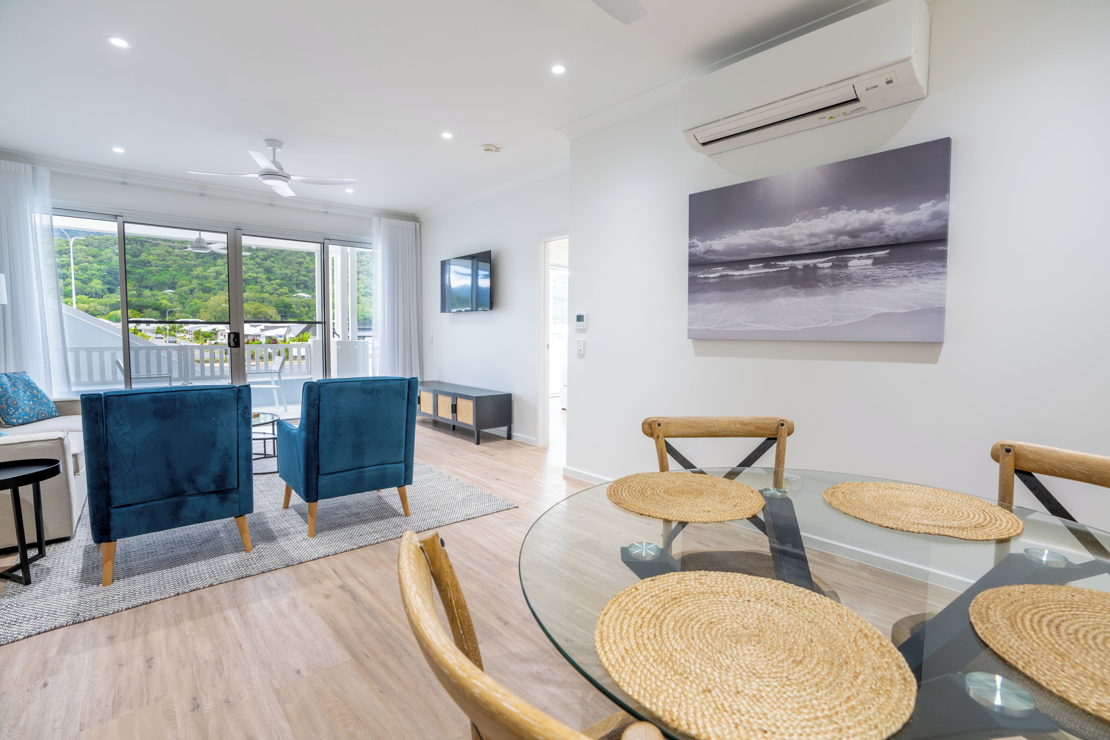 Een moderne woonkamer met een glazen tafel, geweven placemats en uitzicht op een weelderig groen landschap door grote ramen.