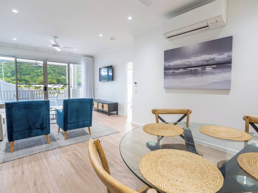 A modern living room with a glass table, woven placemats, and a view of a lush green landscape through large windows.