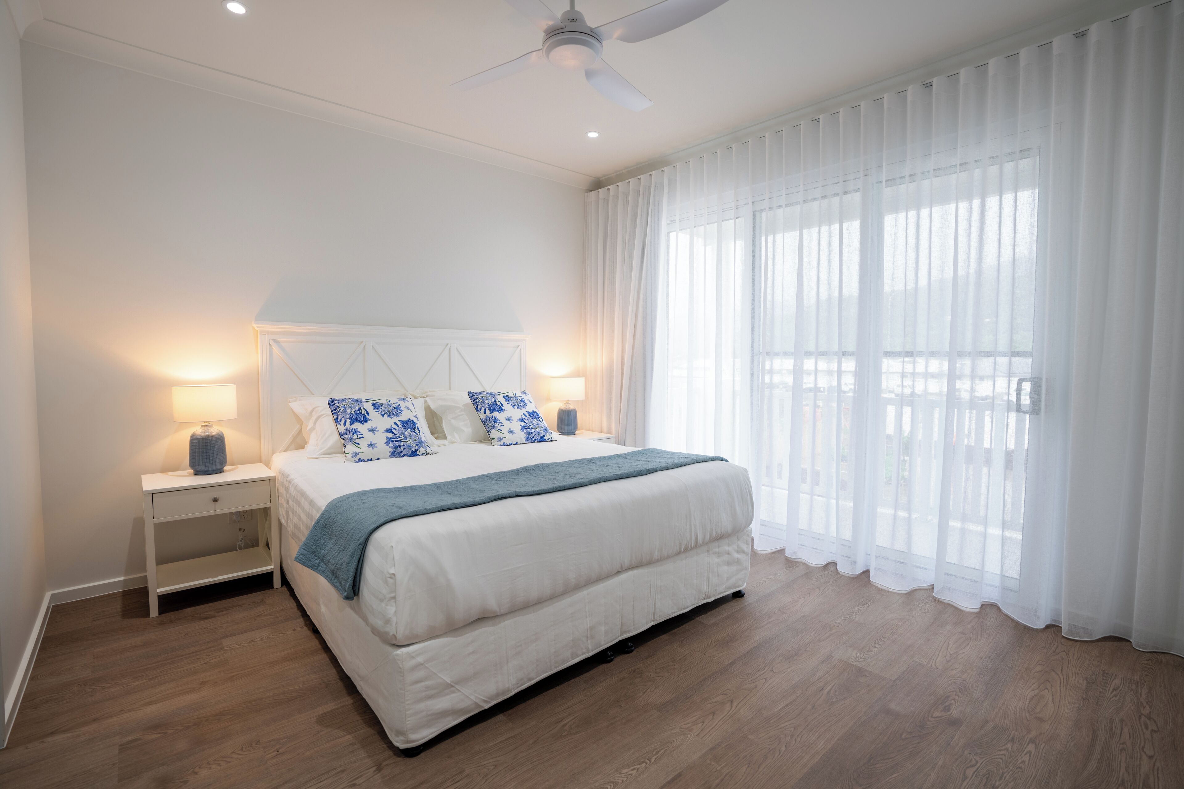 Un dormitorio con una cama grande, cortinas blancas, ventilador de techo y una mesita de noche con lámpara.