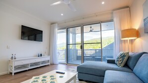 Ein Wohnzimmer mit einer Couch, einem Couchtisch mit sternförmigen Objekten, einem Fernseher an der Wand und einer Glastür, die auf eine Terrasse im Freien führt.