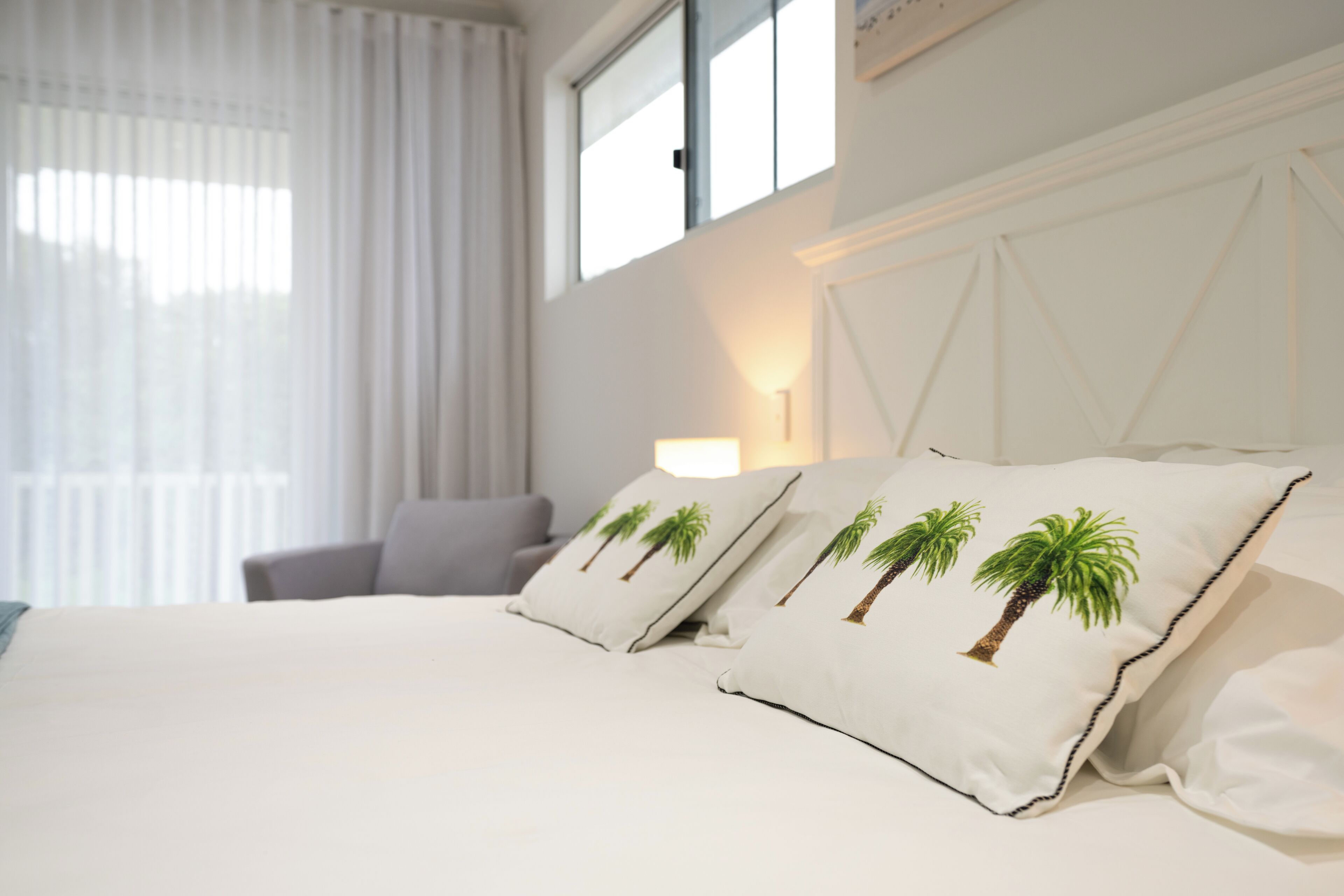 Habitación de hotel con cama, dos almohadas con diseño de palmeras, mesita de noche, lámpara y ventana con cortinas.