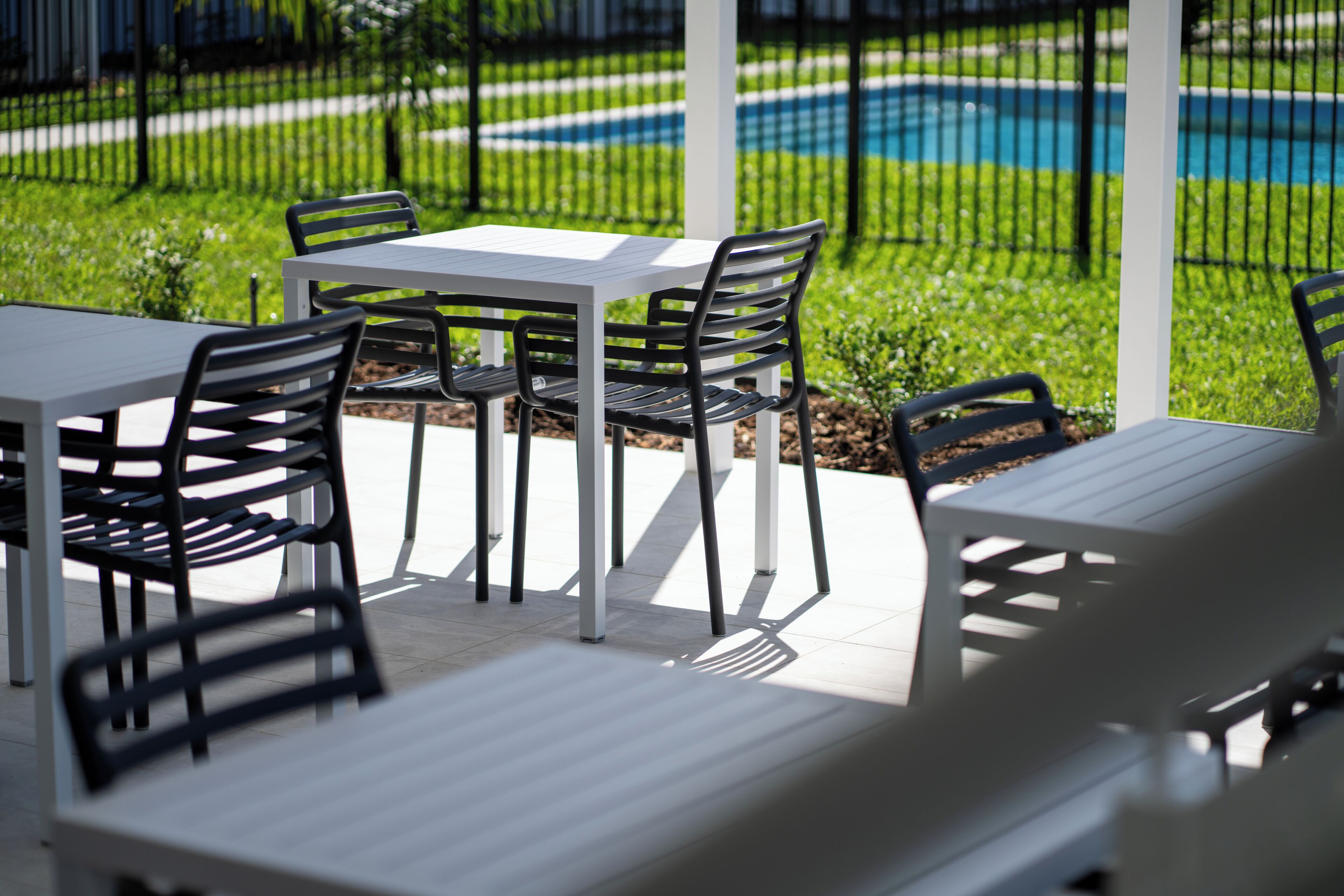 Een terras met een witte tafel en zwarte stoelen, een zwembad op de achtergrond en een groen gazon.