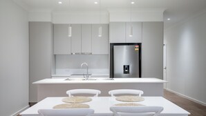 Eine moderne Küche mit weißen Schränken, einem Edelstahlkühlschrank und einer zentralen Kochinsel mit Spüle. Zwei weiße Stühle mit gewebten Platzsets sind vorhanden.