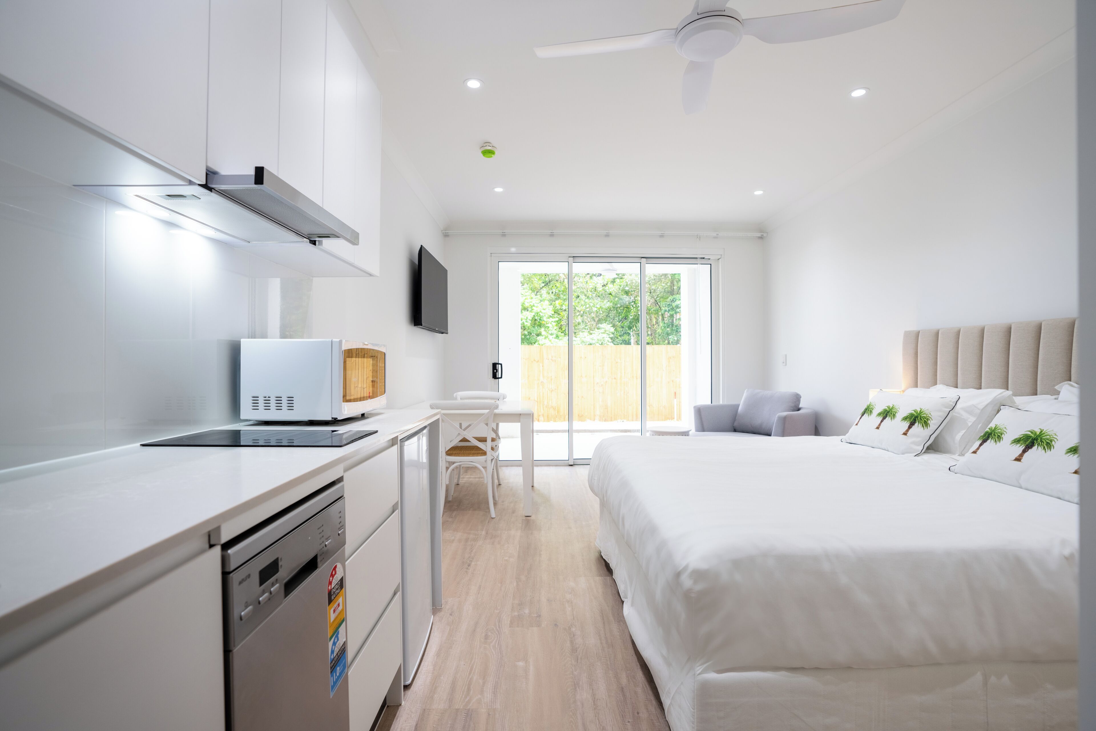 Een moderne hotelkamer met kitchenette, een bed met wit beddengoed en palmboom-kussens, een wit bureau en een groot raam met uitzicht op groen.