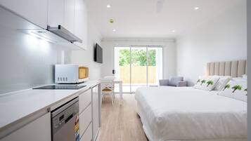 Een moderne hotelkamer met kitchenette, een bed met wit beddengoed en palmboom-kussens, een wit bureau en een groot raam met uitzicht op groen.