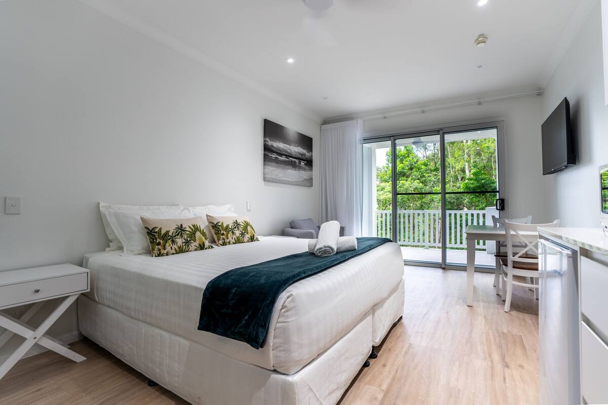 A modern bedroom with a large bed, a desk, and a balcony with a view of greenery.