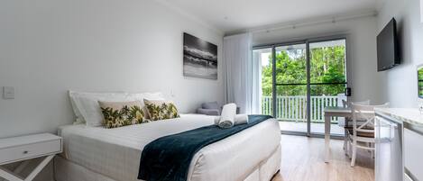 A modern bedroom with a large bed, a desk, and a balcony with a view of greenery.