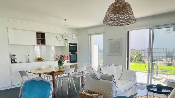 A modern kitchen with white cabinetry, a dining table with chairs, a sofa with white cushions, and a large window offering a view of the outdoors. - Le Sophia à La Baule (La Baule-Escoublac)
