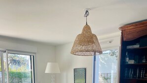 A living room with a sofa, a blue chair, a wicker basket, a round ottoman, a table with a vase of flowers, and a hanging light fixture. - Le Sophia à La Baule (La Baule-Escoublac)