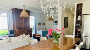A dining area with a long wooden table, white chairs, a chandelier, and a view of the outdoors. - Le Sophia à La Baule (La Baule-Escoublac)