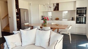 A modern kitchen with white cabinetry, a dining area with a wooden table and chairs, and a living area with a sofa and cushions. - Le Sophia à La Baule (La Baule-Escoublac)