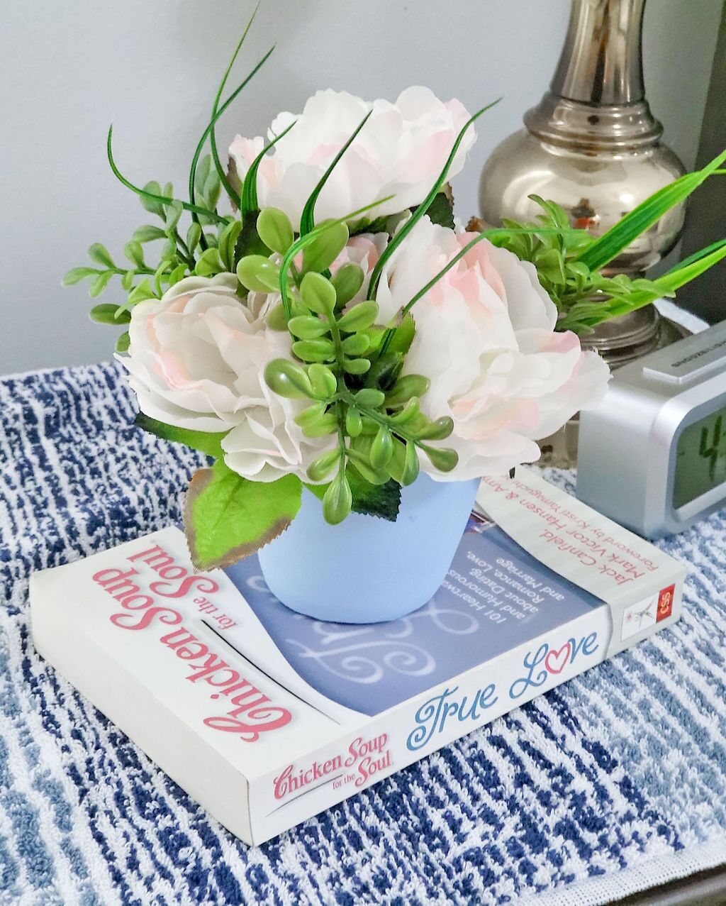 Una planta en test, un mantell de taula estampat amb dibuixos blaus i blancs, un llibre amb el títol 'Chicken Soup for the Soul' i un rellotge despertador platejat.