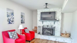 A living room with a stone fireplace, two red armchairs, a TV mounted above the fireplace, and a wooden floor. - Amazing Women’s and Men’s Masters Tournament Rental- Fully Remodeled. (Martinez)