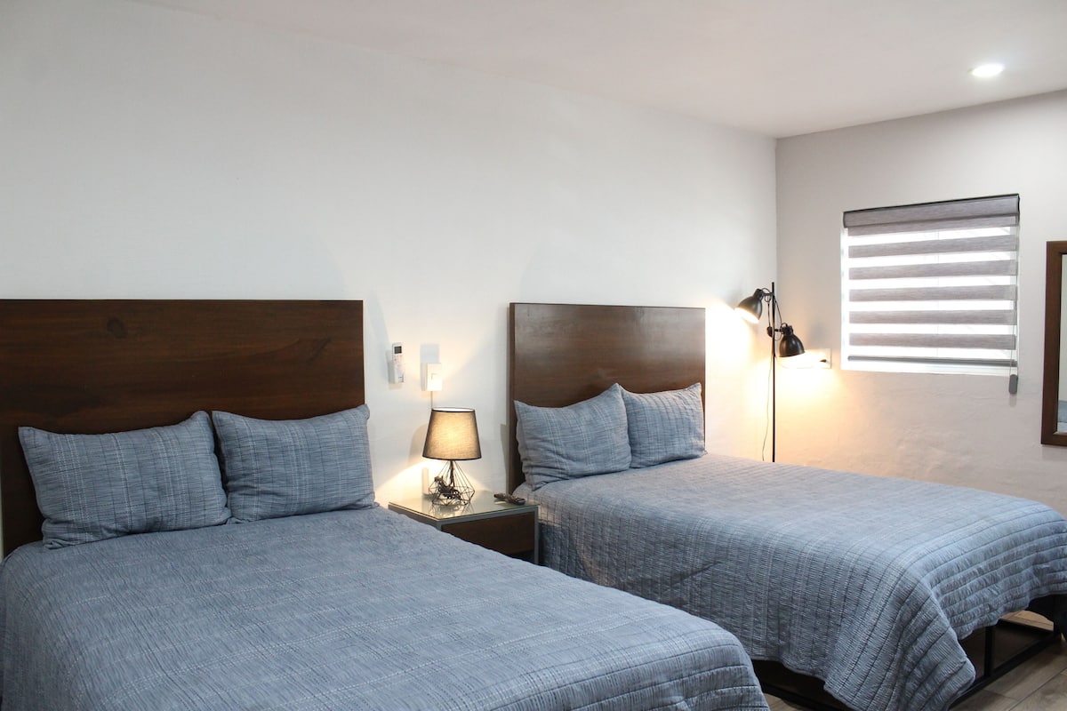 A hotel room with two beds, a nightstand with a lamp, and a window with blinds.