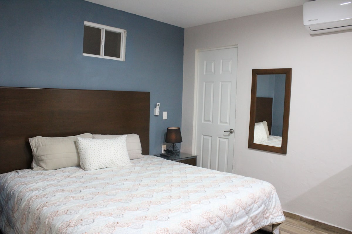 A bedroom with a bed, a nightstand, a wall-mounted lamp, a door, and a window.