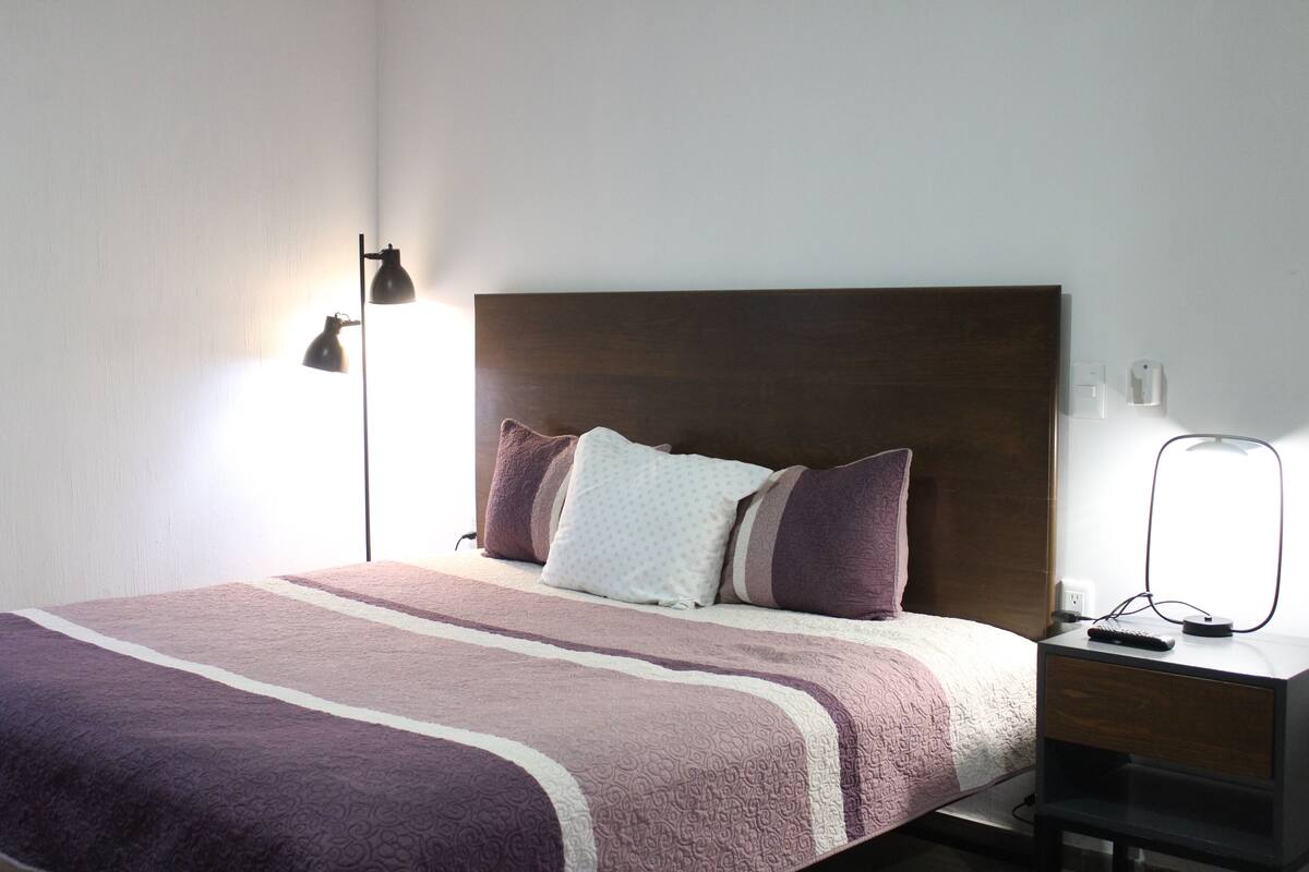 A neatly made bed with a patterned bedspread, a dark headboard, and a bedside table with a lamp and remote control.