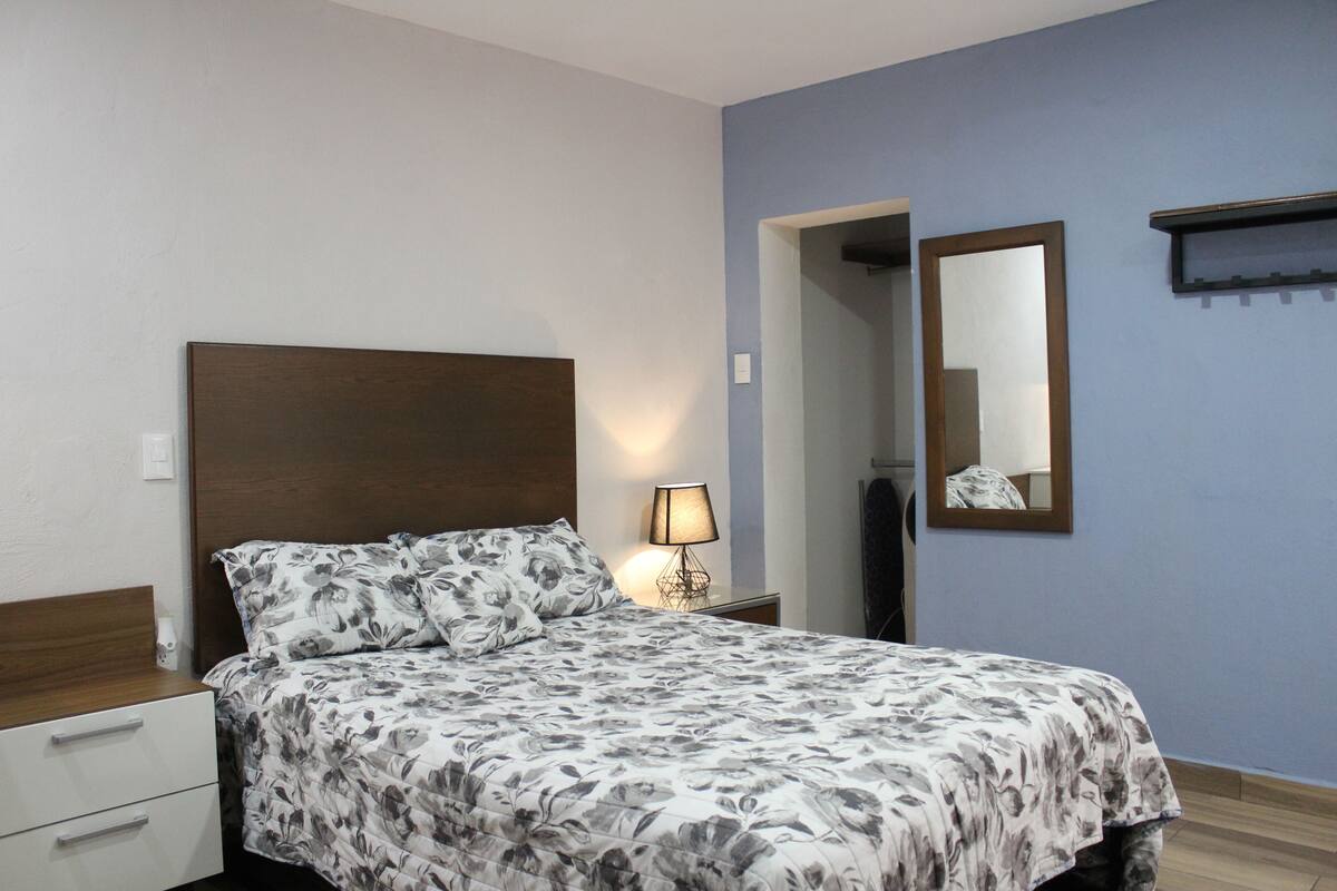 A bedroom with a bed, a bedside table, a lamp, a mirror, and a wooden nightstand.