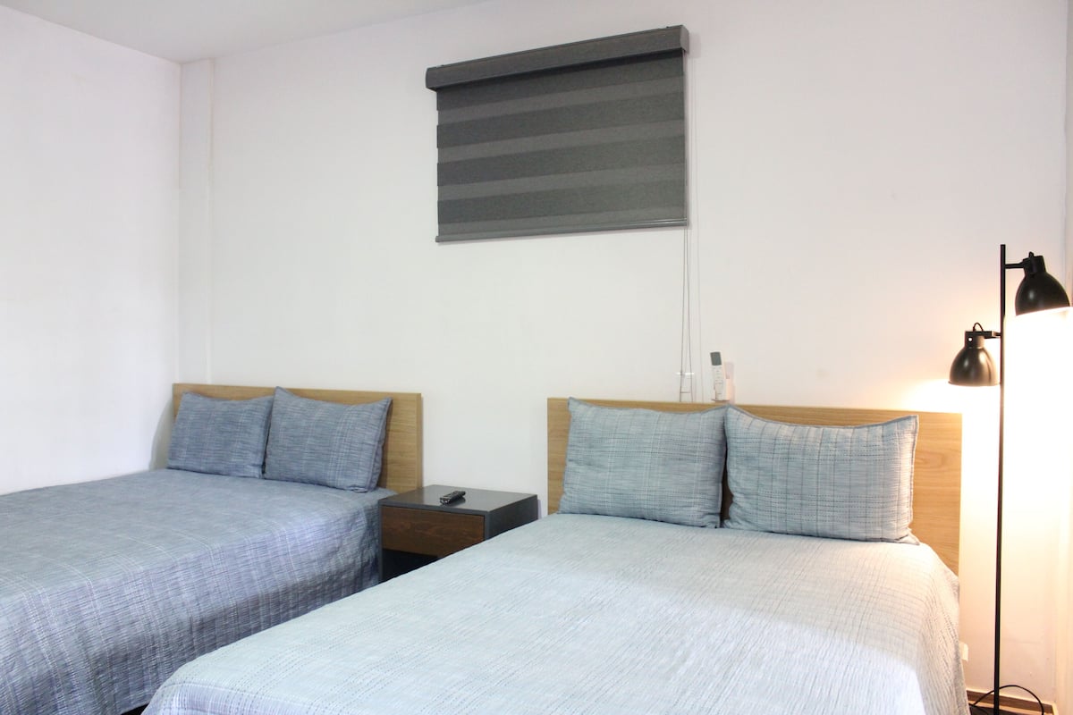 A room with two beds, a nightstand, a lamp, and a window with blinds.
