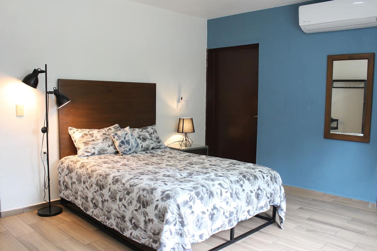 A bedroom with a bed, bedside lamps, a nightstand, a wooden headboard, a door, and an air conditioning unit.