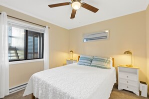 A bedroom with a bed, two bedside tables with lamps, a ceiling fan, a window with blinds, and a painting on the wall.