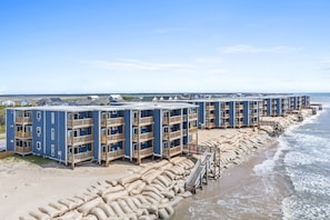 Un hotel frente a la playa con edificios de varios pisos, balcones de madera y una playa de arena en primer plano.