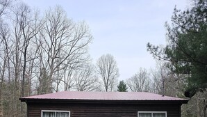 Une cabane en bois avec un toit en métal, deux fenêtres avec des rideaux et une véranda avec deux chaises.