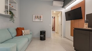 A modern living room with a light blue sofa, a small black trash bin, a flat-screen TV mounted on the wall, and a kitchen area with a dark cabinet and a knife block. - Champagne Loft (Milan)