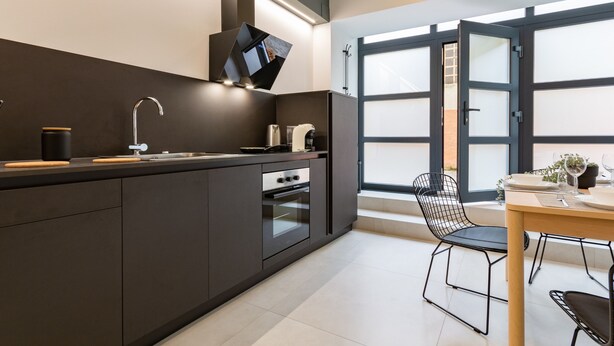 A modern kitchen with dark cabinetry, a stainless steel sink, and a built-in oven. There is a dining area with a table and chairs, and a window with frosted glass panels. - Champagne Loft (Milan)