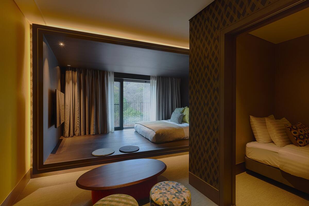 A modern hotel room with a large window, a bed, a seating area with a table and chairs, and a patterned wall.