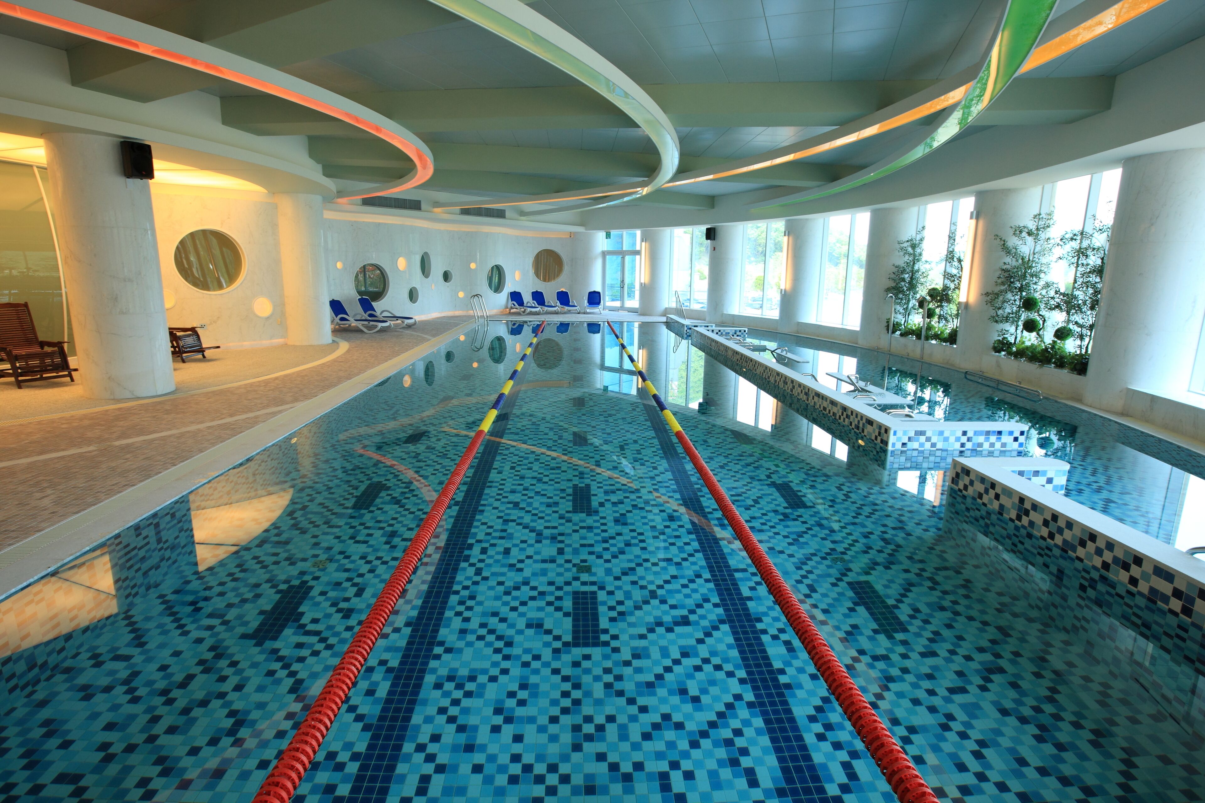 Piscina interior con carriles delimitados, iluminada por amplios ventanales y rodeada de sillones y bancos.