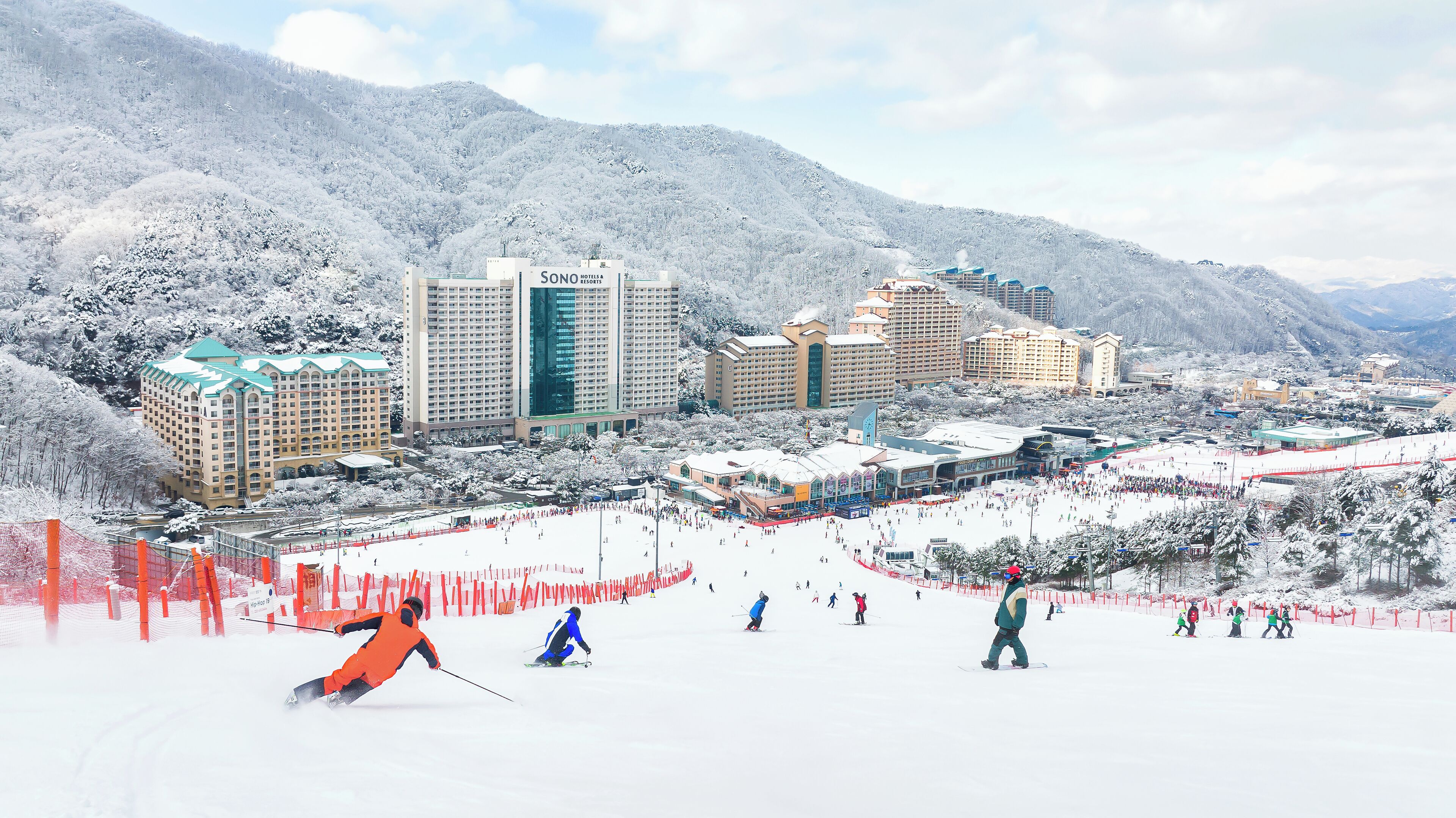 Ein Skigebiet mit mehreren Gebäuden, darunter ein Hotel namens SONO, und einer Skipiste mit Skifahrern und Snowboardern.