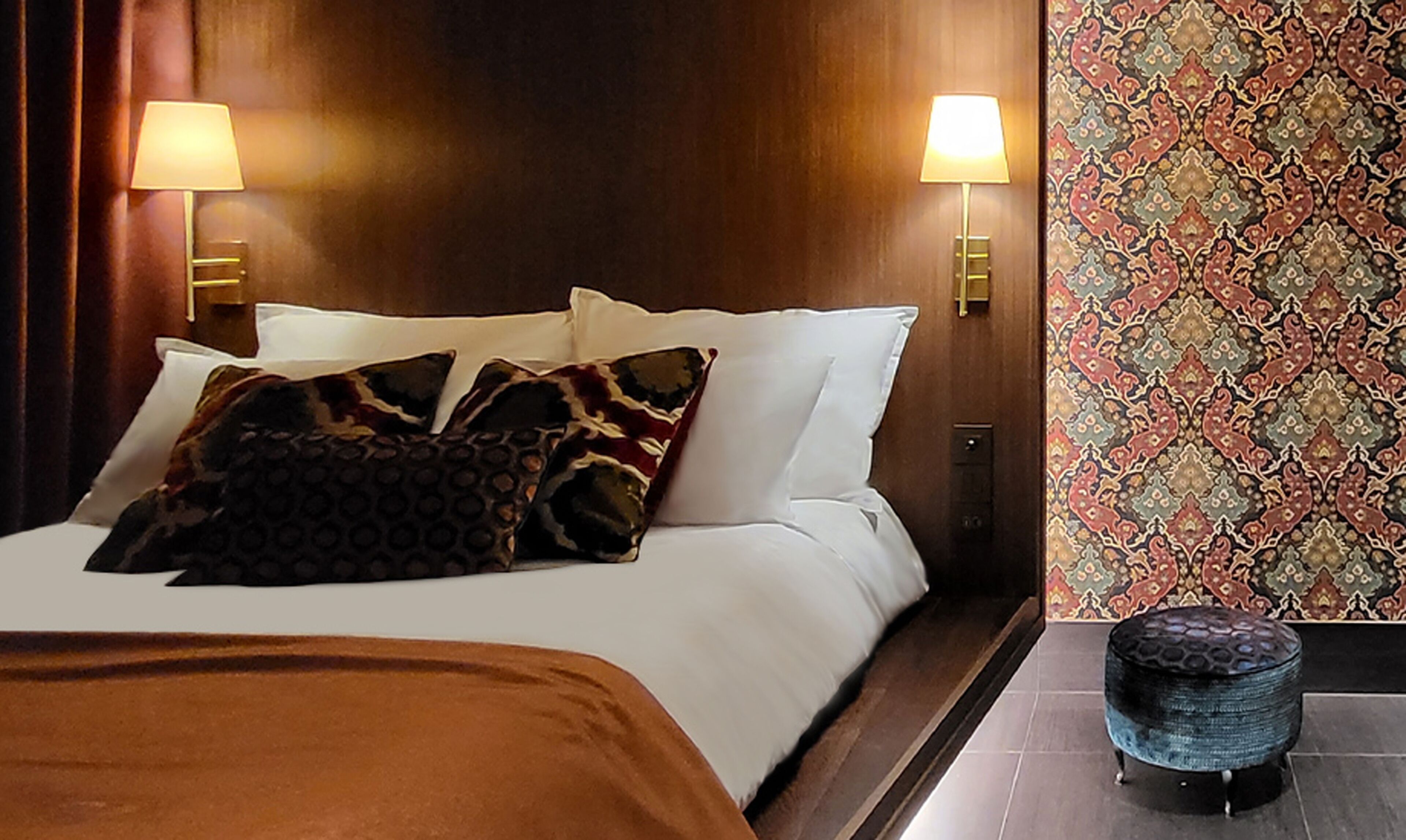Habitación de hotel con cama, dos lámparas de pared, pared con diseño y un pequeño otoman.