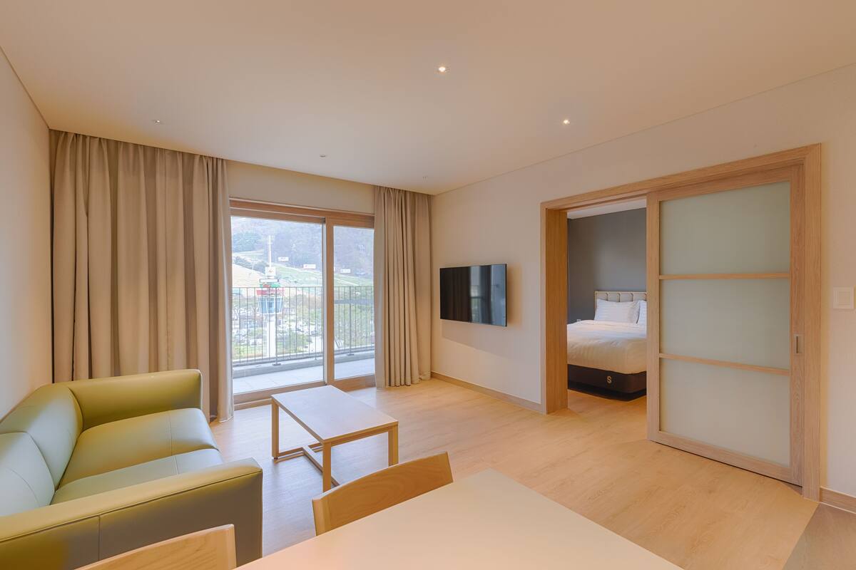 A modern hotel room with a sofa, a small table, a bed with a sliding door, a TV, and a balcony with curtains.