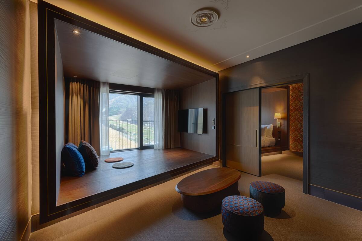 A modern hotel room with a large sliding glass door leading to a balcony, a flat-screen TV, a wooden coffee table, and two patterned ottomans.