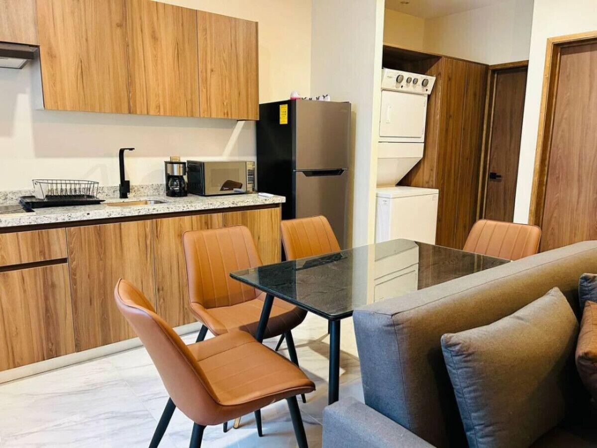 Une cuisine moderne avec des meubles en bois, un réfrigérateur en acier inoxydable, un four à micro-ondes et une machine à café. On y trouve également un coin repas avec une table en verre et des chaises en cuir.