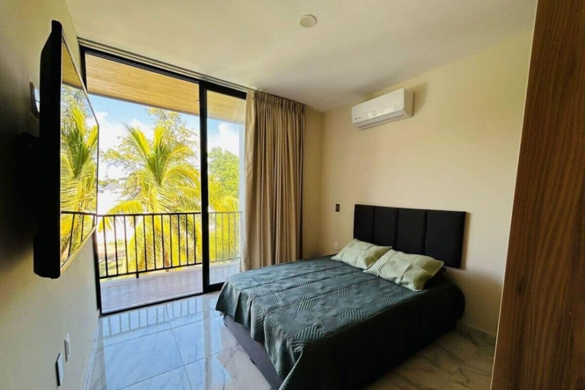A bedroom with a bed, a TV mounted on the wall, and a balcony with a view of palm trees.