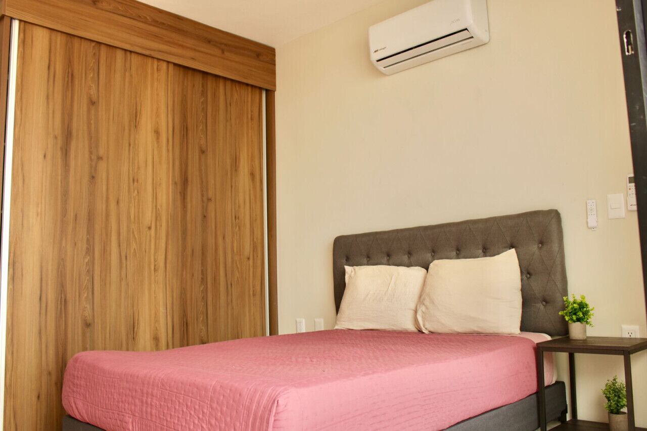 A bedroom with a bed, a wooden wardrobe, a grey upholstered headboard, a bedside table with a plant, and an air conditioning unit on the wall.