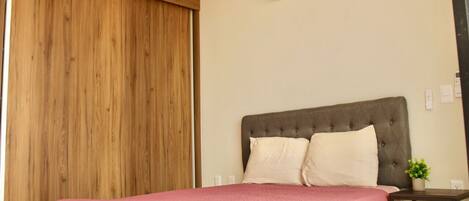 A bedroom with a bed, a wooden wardrobe, a grey upholstered headboard, a bedside table with a plant, and an air conditioning unit on the wall.