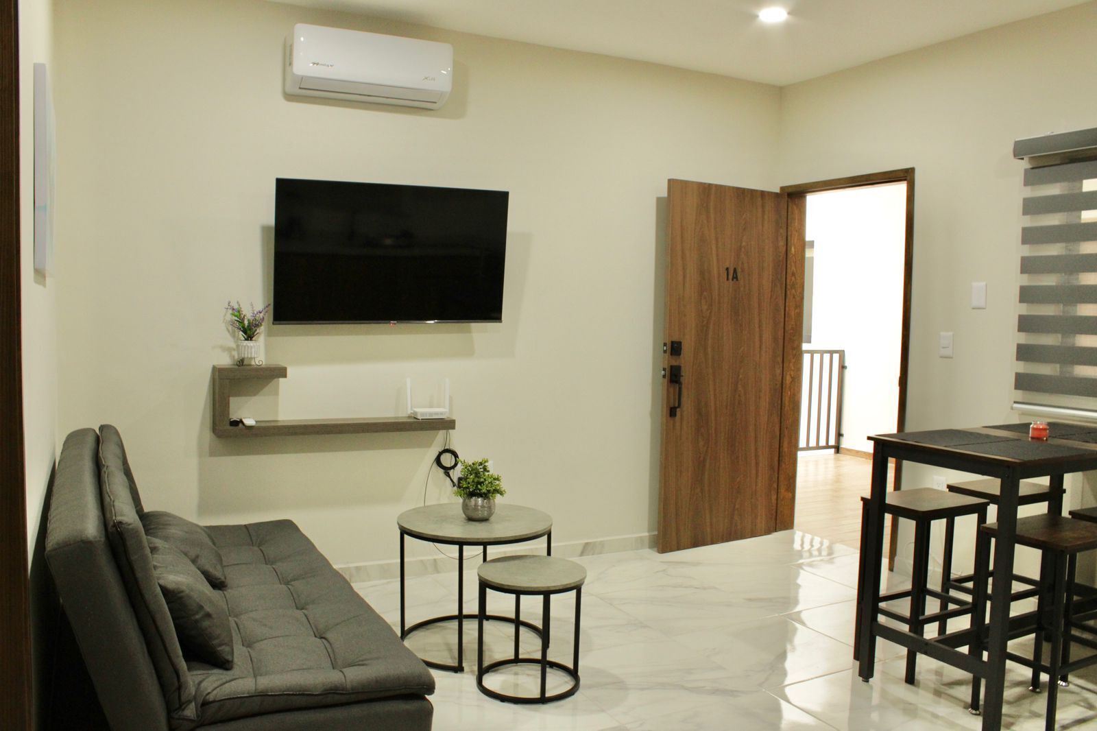A modern living room with a flat-screen TV, a grey sofa, a small round table with a plant, and a wooden door leading to another room.