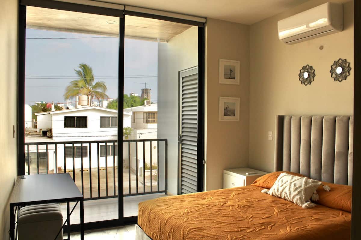 A bedroom with a bed, a desk, a chair, a balcony, and an air conditioning unit.