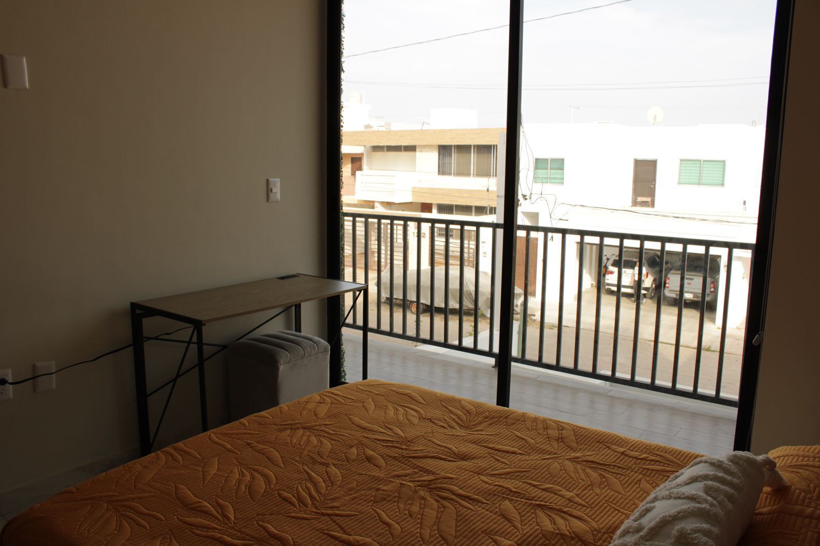 A room with a bed, a desk, and a view of a street through a balcony.