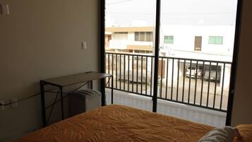 A room with a bed, a desk, and a view of a street through a balcony.