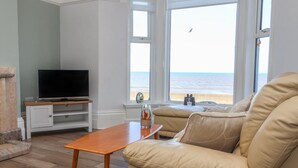 Un salon avec un canapé beige, une table basse en bois, un téléviseur posé sur un meuble blanc et une vue sur la mer grâce à de grandes fenêtres.